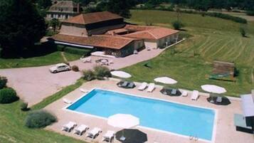 A large rectangular swimming pool with lounge chairs and umbrellas, surrounded by a well-maintained lawn and a large building with a tiled roof.