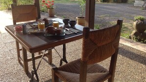 Ein Holztisch mit Frühstücksartikeln, darunter Croissants und Orangensaft, draußen mit Blick auf ein Gebäude und Grün im Hintergrund.