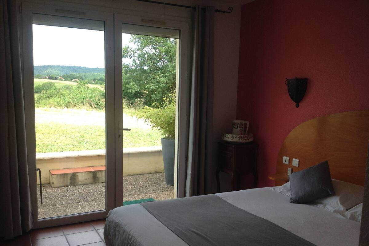Habitación de hotel con una cama, una mesita con un objeto decorativo y vistas a un paisaje verde a través de una puerta corrediza.