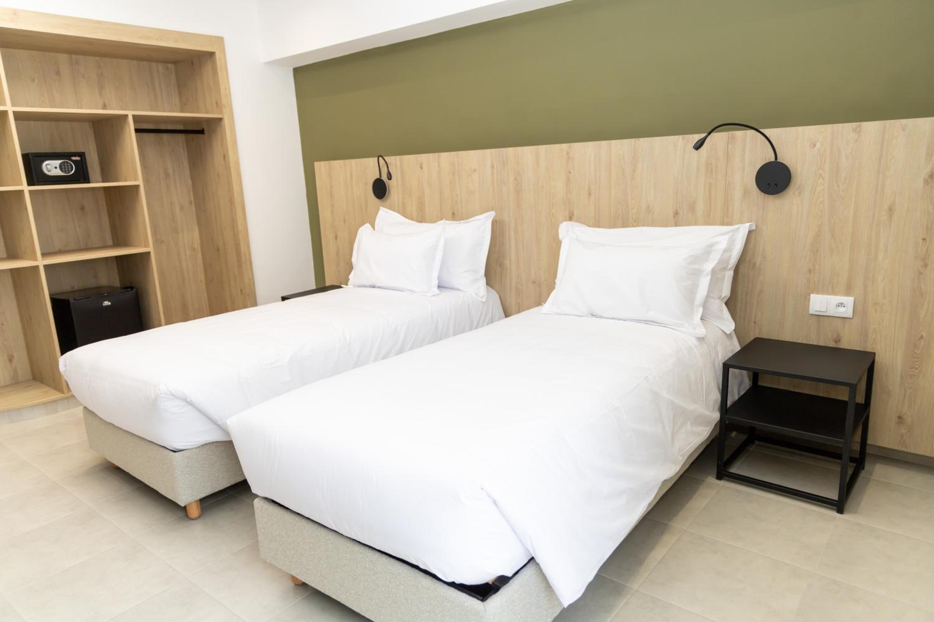 Habitación de hotel con dos camas individuales, cabecero de madera, una mesita de noche pequeña y una estantería con un radio.