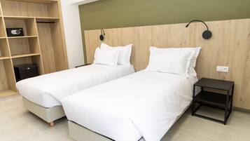 Habitación de hotel con dos camas individuales, cabecero de madera, una mesita de noche pequeña y una estantería con un radio.