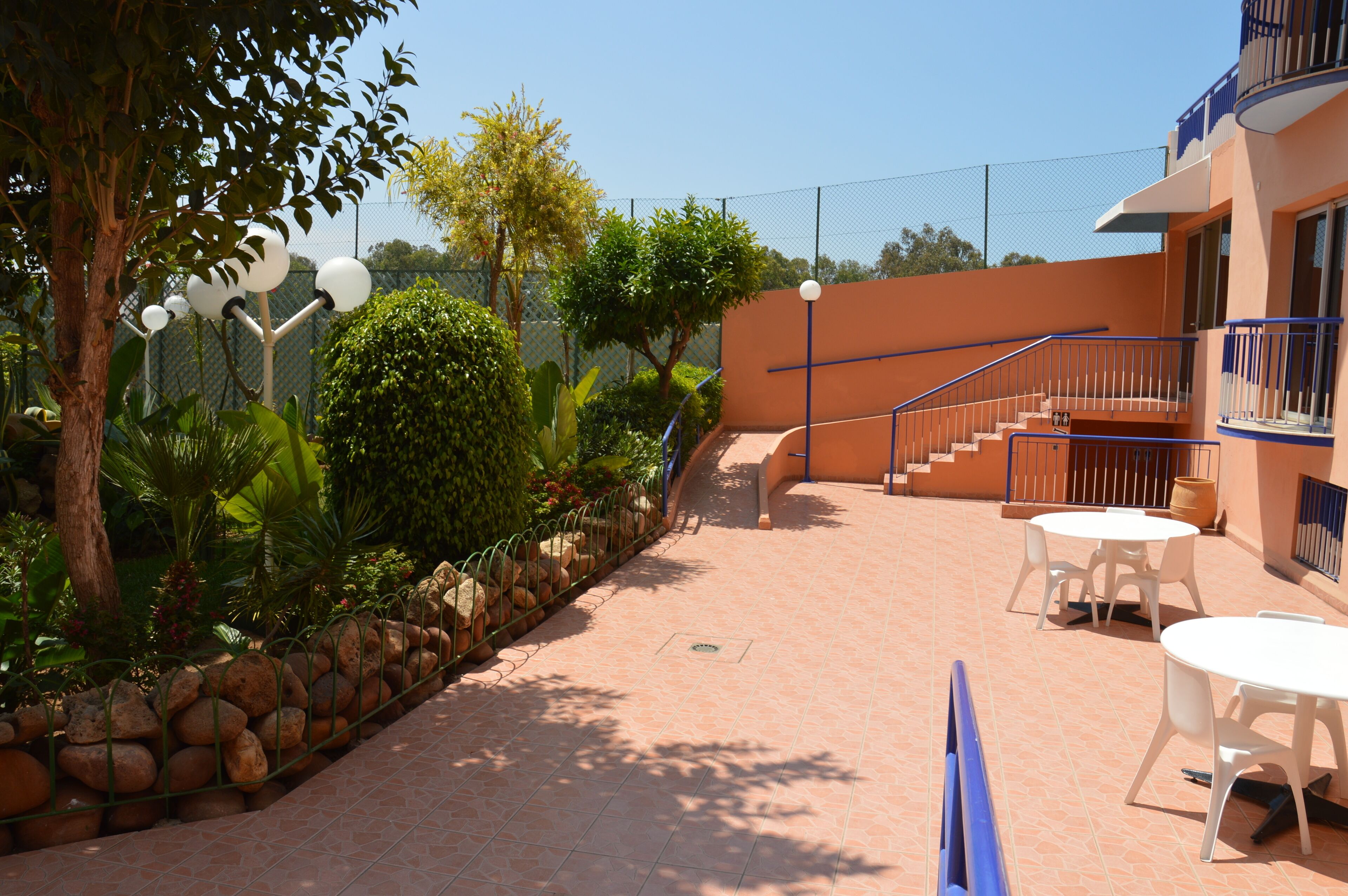 A patio area with a tiled floor, white tables and chairs, a stone wall, and a staircase leading to an upper level.