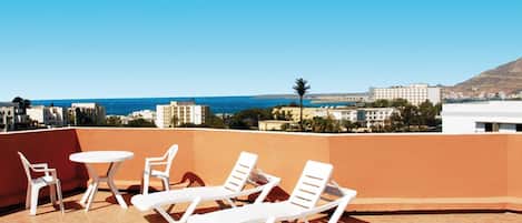 A rooftop terrace with white lounge chairs and a small table, overlooking a coastal cityscape with buildings and a clear blue sky.