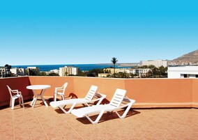 Terraza en la azotea con sillones blancos y una pequeña mesa, con vista a un paisaje urbano costero con edificios y un cielo azul despejado.