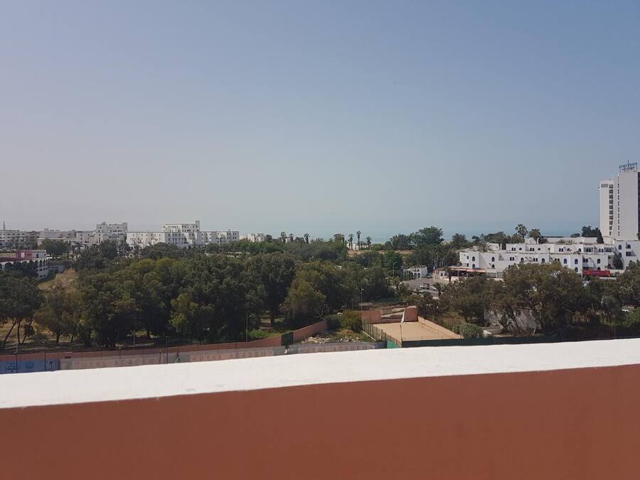 Un paisaje urbano costero con un edificio blanco destacado y la inscripción “ROYAL ACADEMY” a la vista, cielo despejado y vista al mar en la distancia.