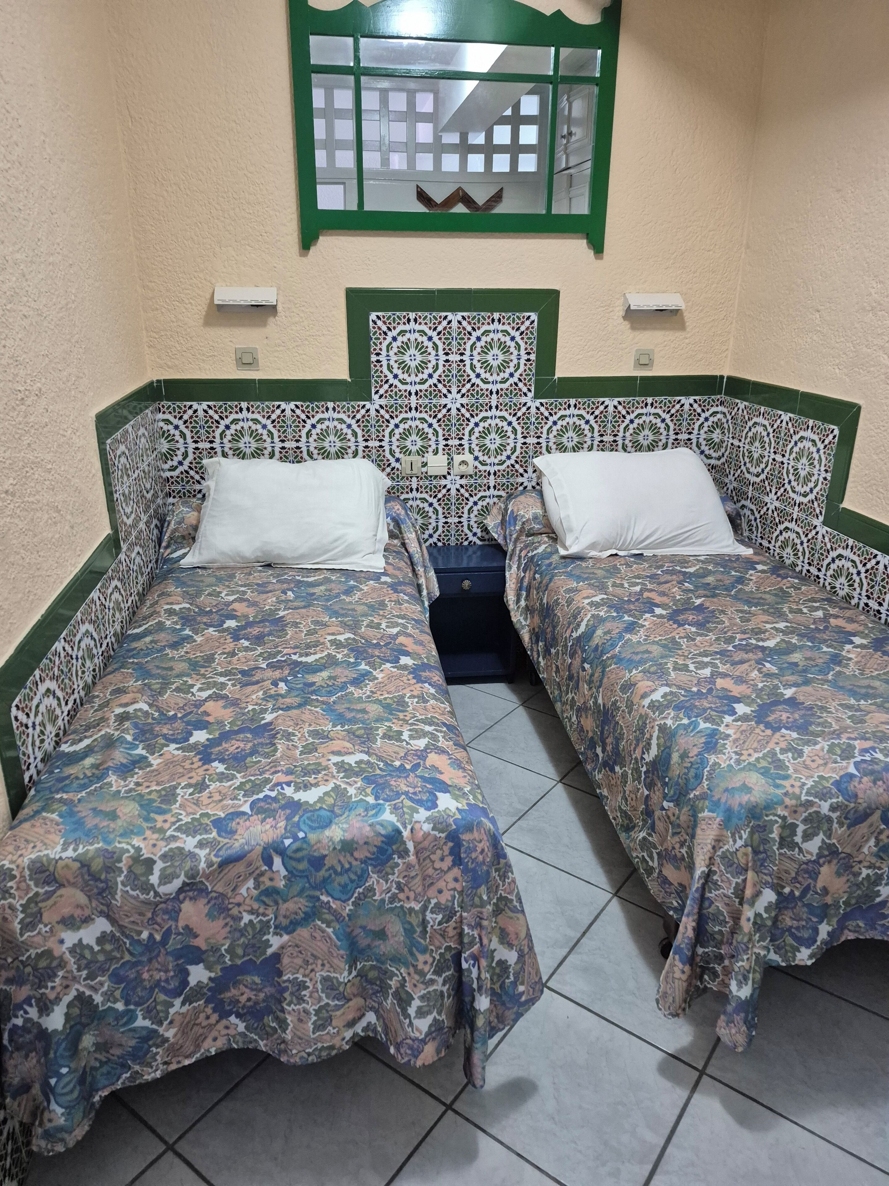 Une chambre avec deux lits, chacun recouvert d’une couvre-lit à motifs, un miroir à cadre vert au-dessus, et un sol carrelé.