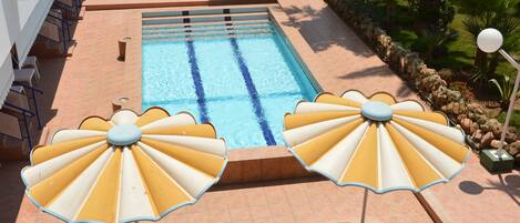 A swimming pool with two sun umbrellas, lounge chairs, and a tiled patio area.