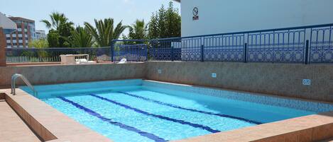 A swimming pool with a tiled deck, a metal railing, and a view of buildings and palm trees in the background.