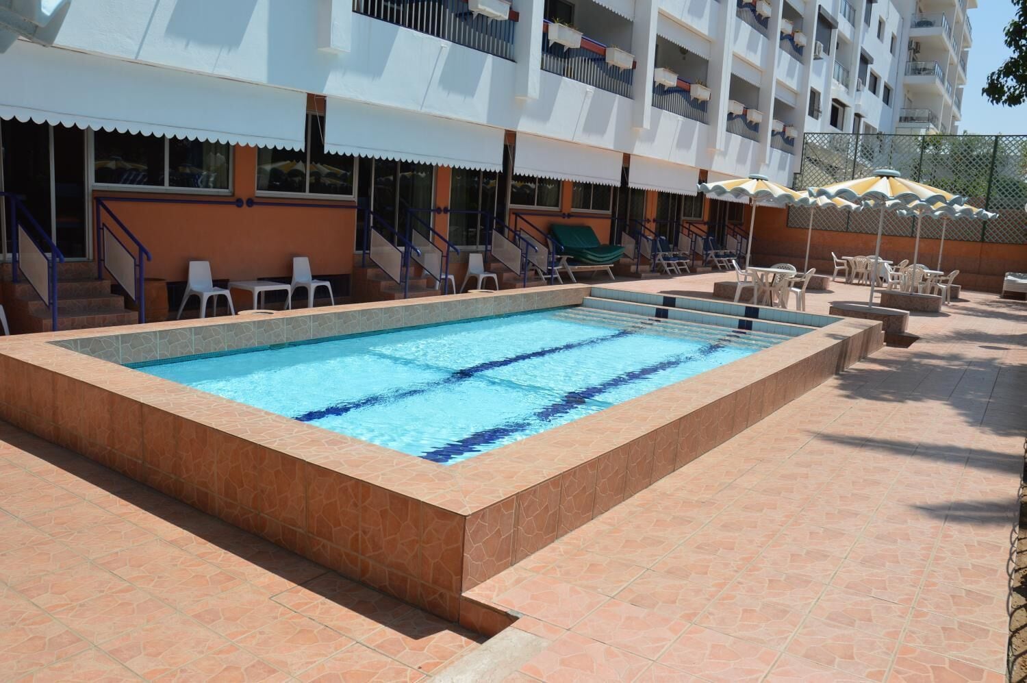 A swimming pool with a tiled deck, surrounded by lounge chairs and umbrellas, and a building with balconies in the background.