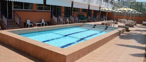 A swimming pool with a tiled deck, surrounded by lounge chairs and umbrellas, and a building with balconies in the background.