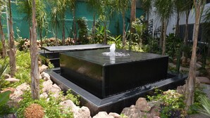 Un bassin rectangulaire noir, agrémenté d’une fontaine, est aménagé dans un jardin, entouré de rochers et de différentes plantes.