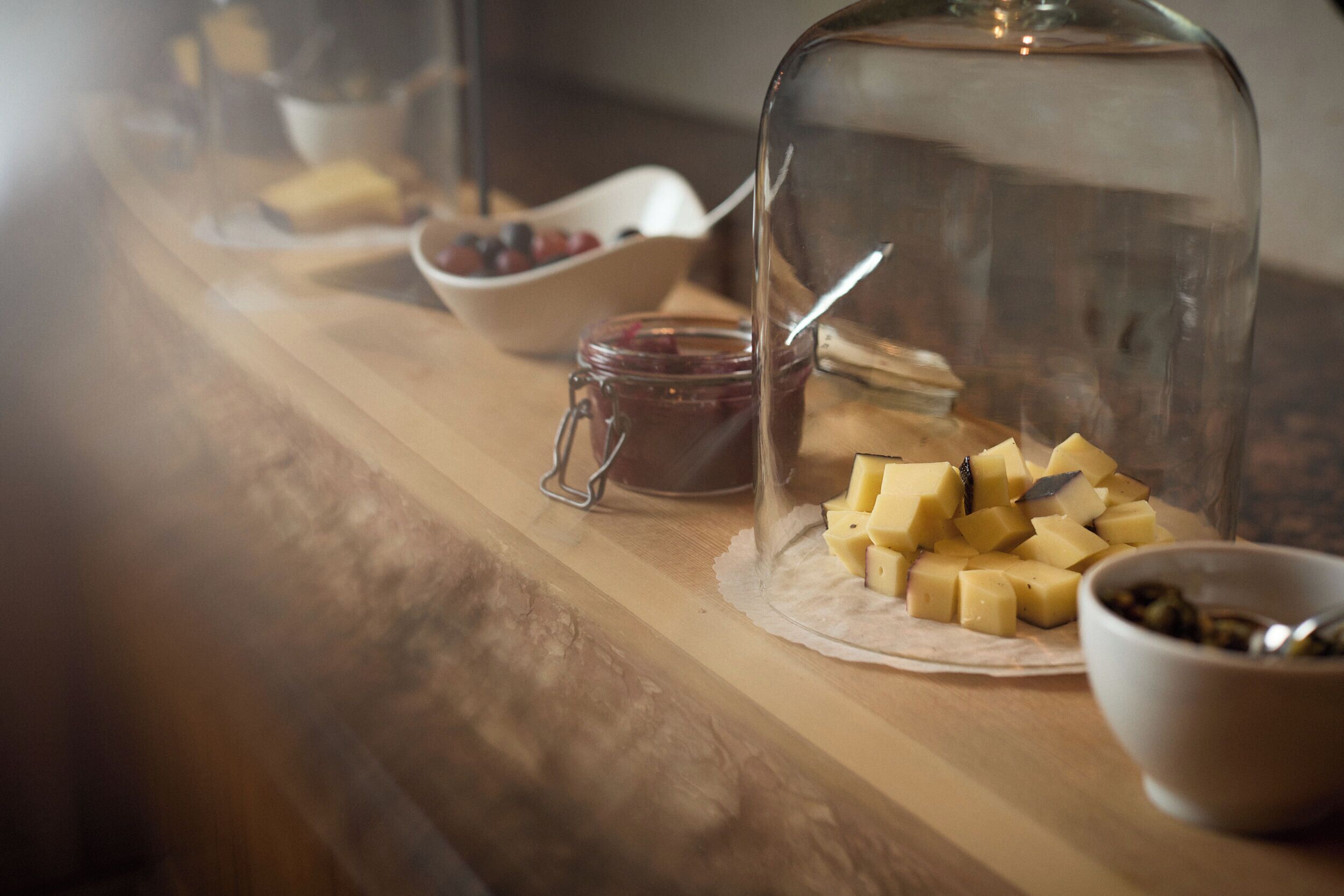 A wooden table with a glass dome cover, a bowl of cheese, a jar of jam, and a bowl of berries.