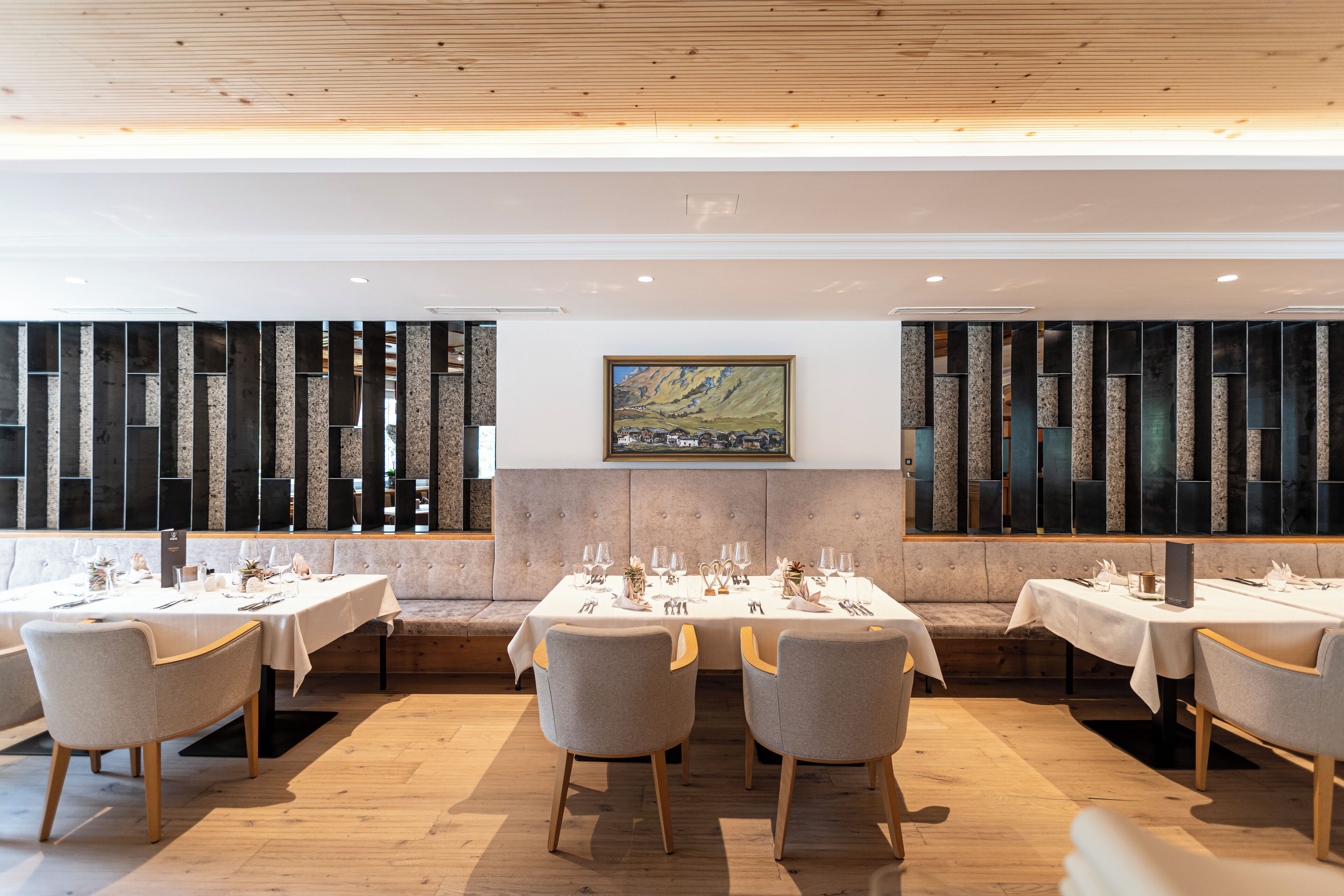 A restaurant with multiple tables set for dining, a cushioned bench seating area, and a large painting on the wall.