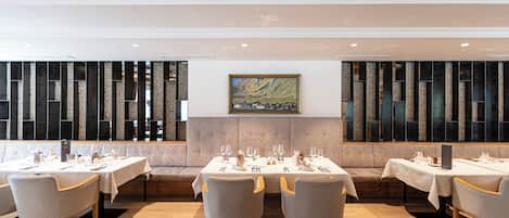 A restaurant with multiple tables set for dining, a cushioned bench seating area, and a large painting on the wall.