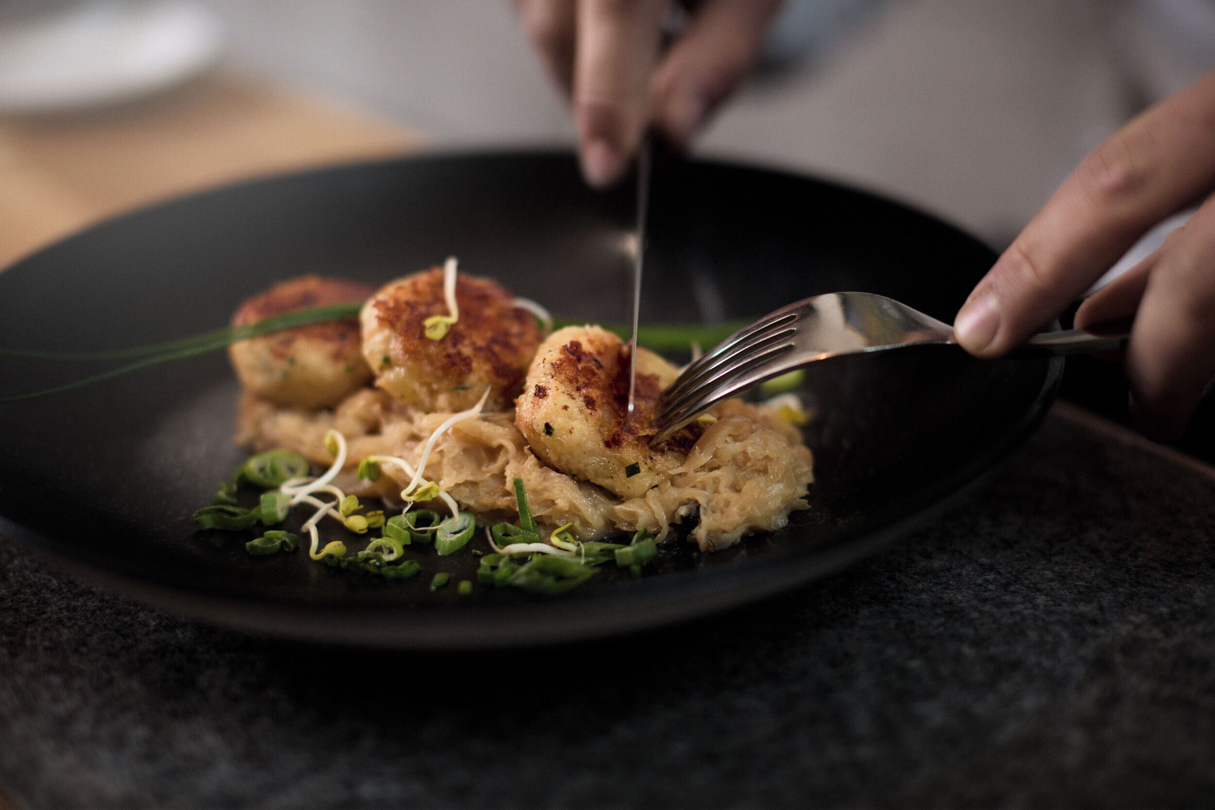 A person using a fork to cut a dish with seared scallops and greens on a black plate.