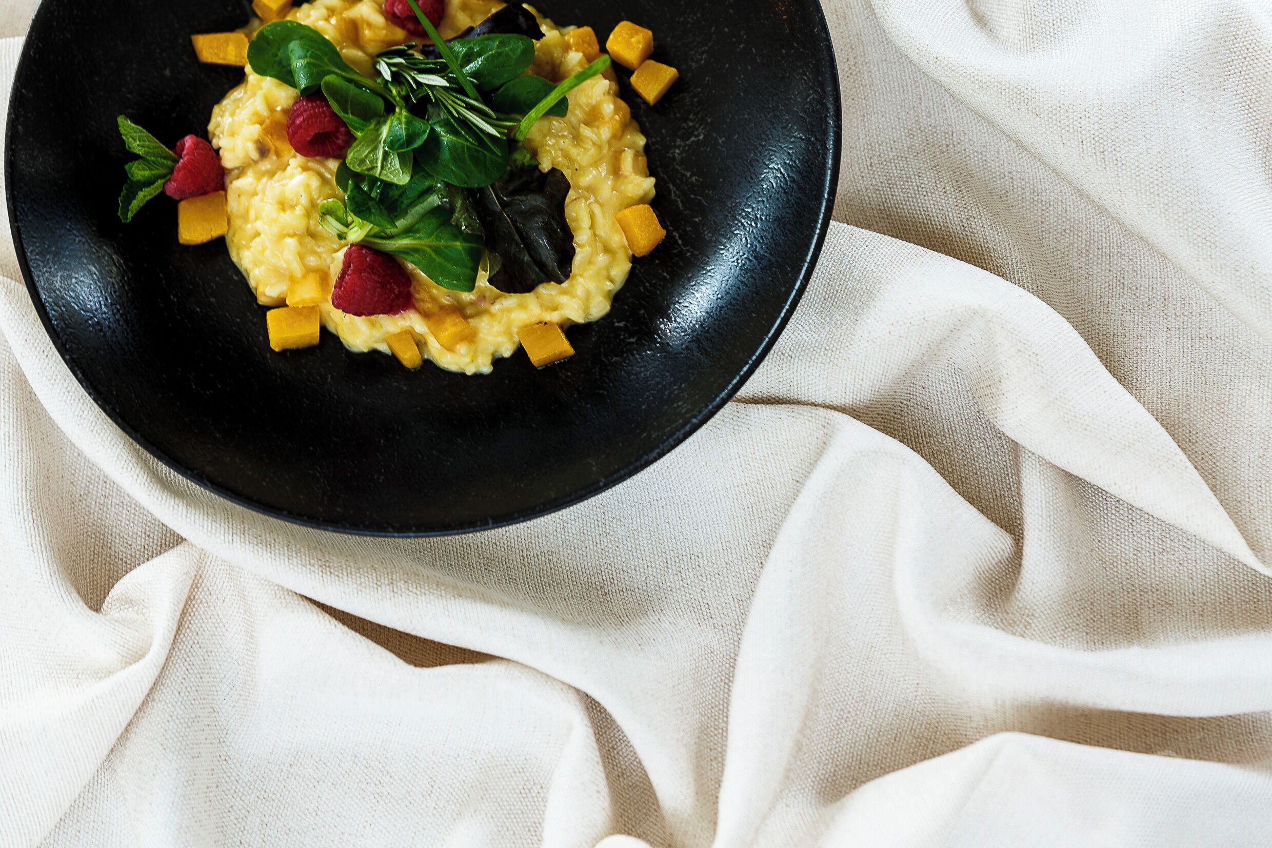 A black plate with scrambled eggs, diced mango, raspberries, and spinach on a white cloth.