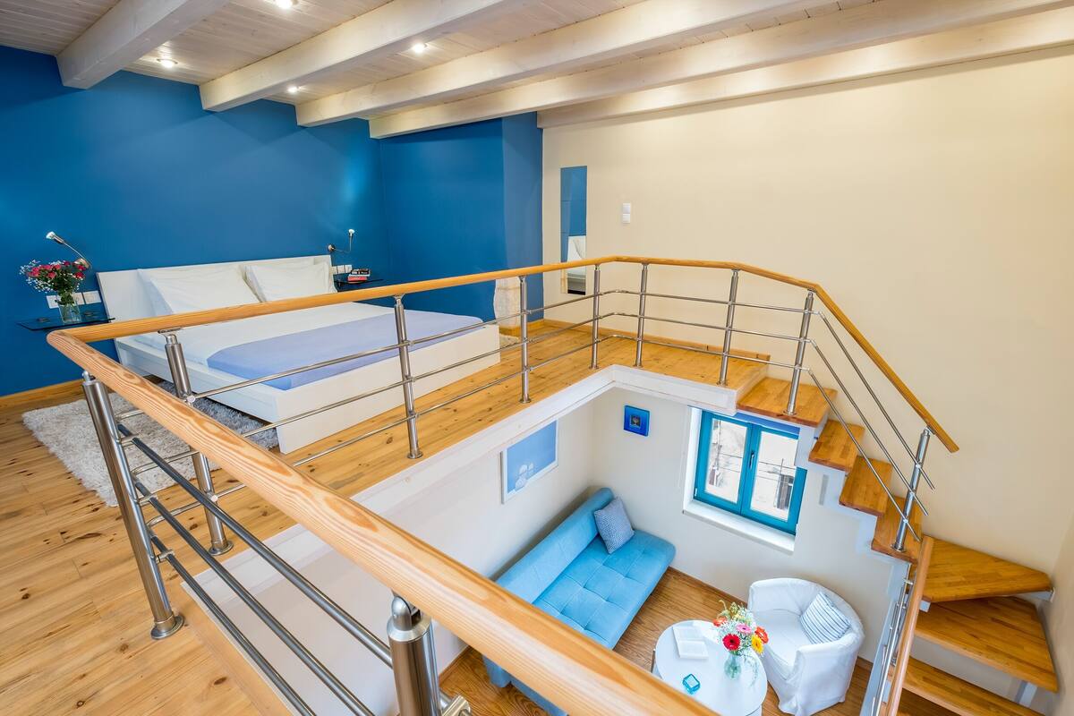 A modern loft bedroom with a wooden staircase, a blue sofa, and a small dining area.