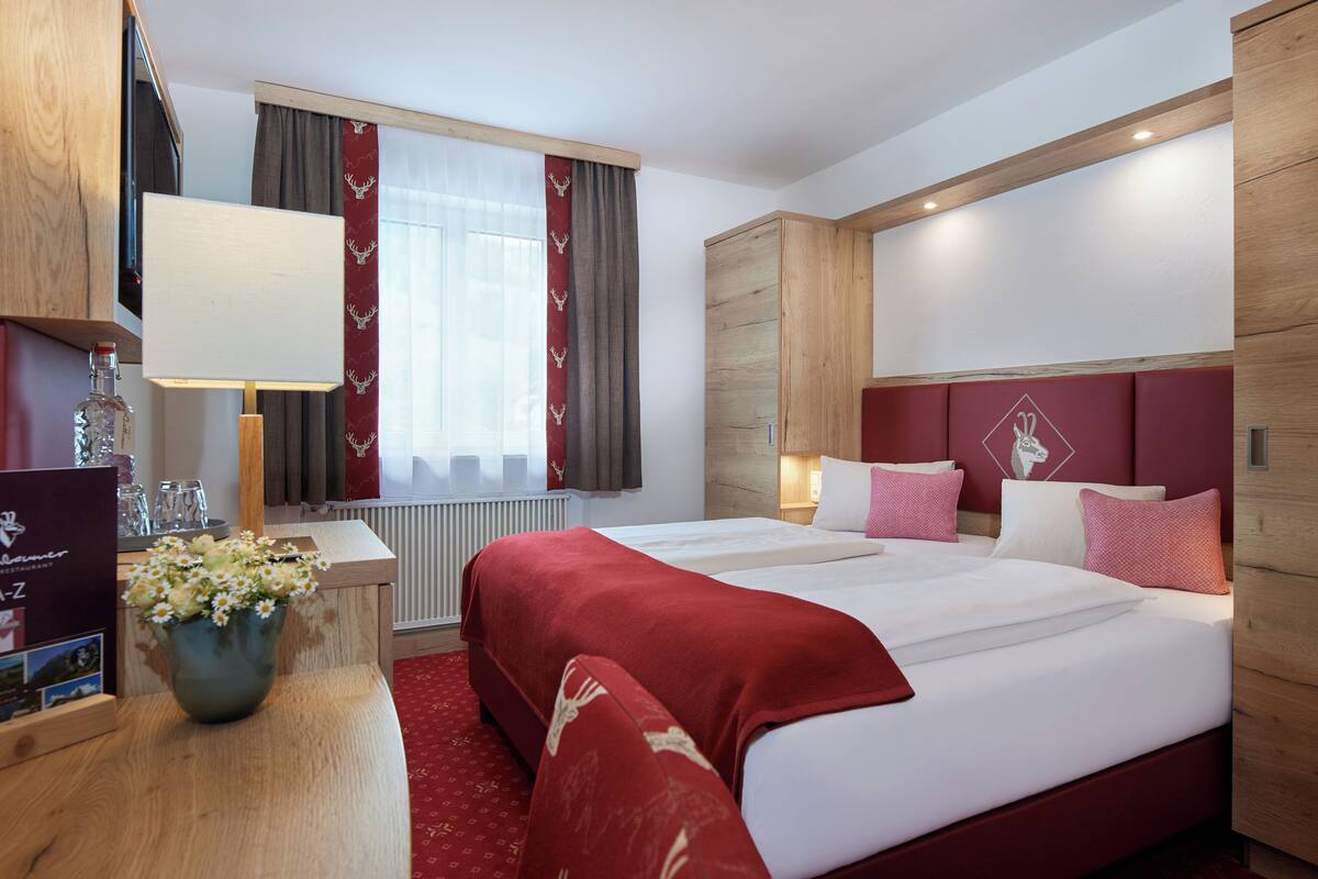 Hotelkamer met twee bedden, een houten hoofdbord, een rode accentmuur, een raam met gordijnen, een nachtkastje met lamp en een bloevas.