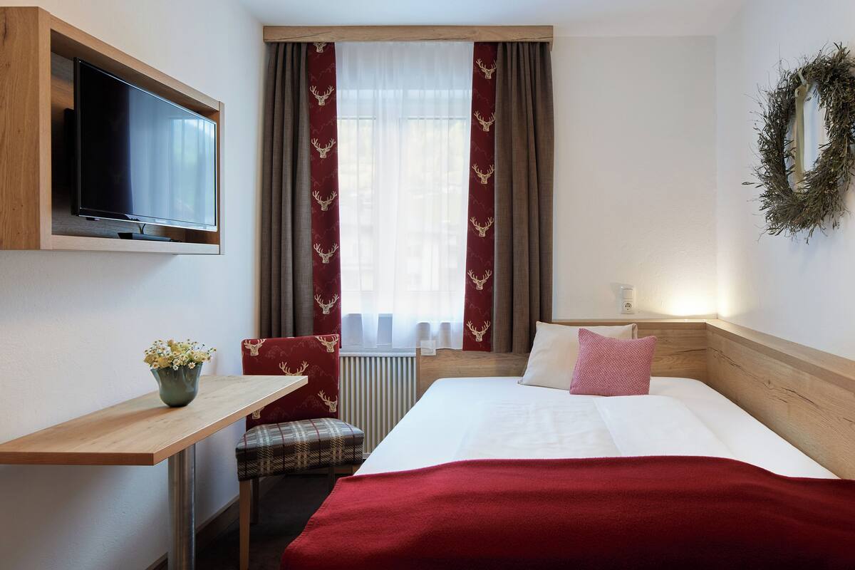 Een hotelkamer met een bed, een aan de muur bevestigde televisie, een kleine tafel met een plant, een geruite fauteuil en een raam met gordijnen.