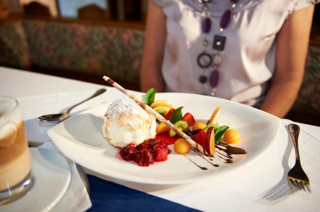 Un piatto dessert con una pallina di gelato, frutta e salsa al cioccolato, accompagnato da forchetta e cucchiaio su un tavolo.