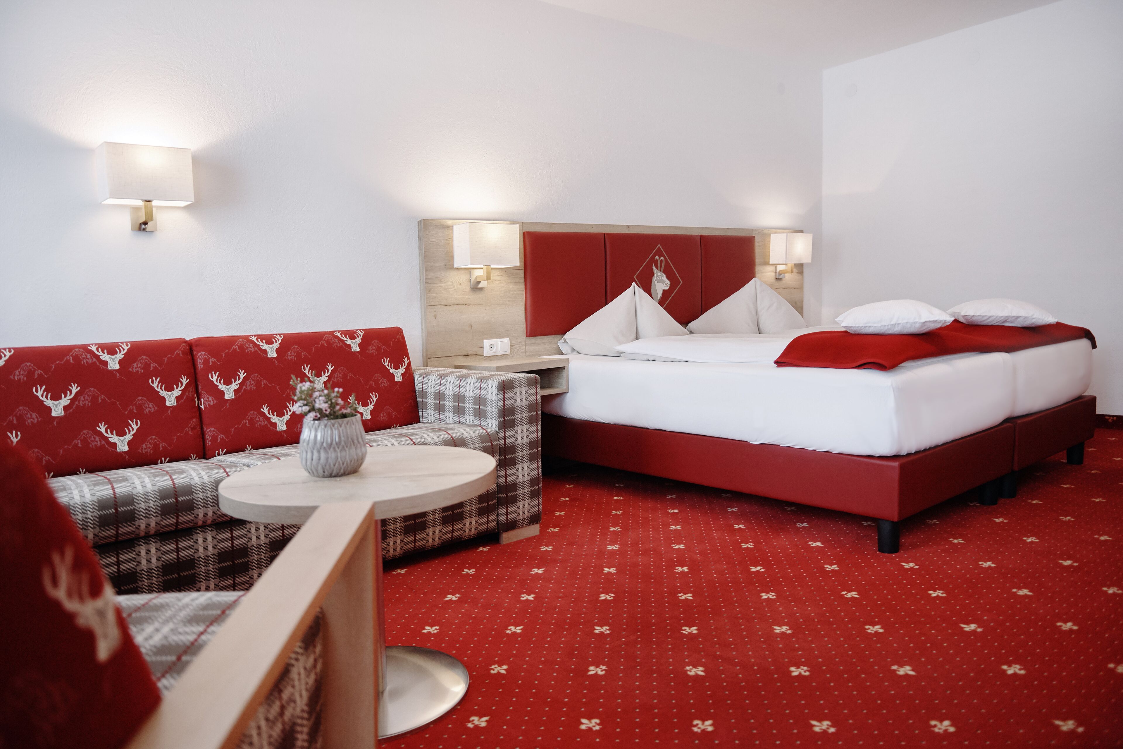 Une chambre d’hôtel aux couleurs rouge et blanc, avec un lit, un canapé, une petite table et deux lampes fixées au mur.