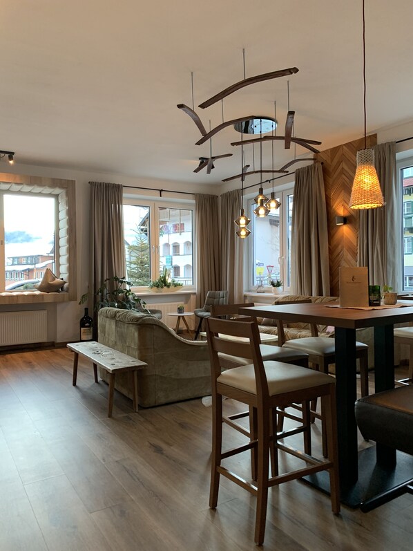 A modern living room with a wooden dining table, cushioned seating, and a large window with curtains.