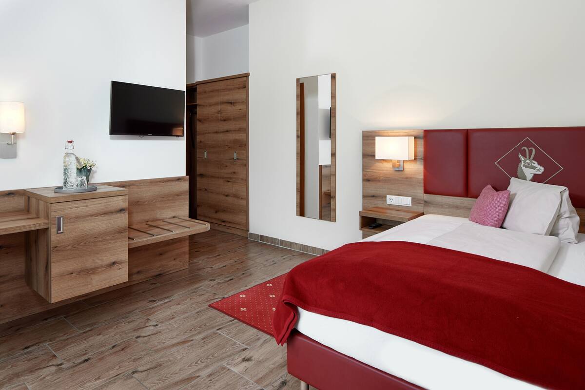 Een moderne hotelkamer met een houten dressoir, een flatscreen televisie, een grote spiegel, een bed met een rode hoofdbord en een nachtkastje met een lamp.