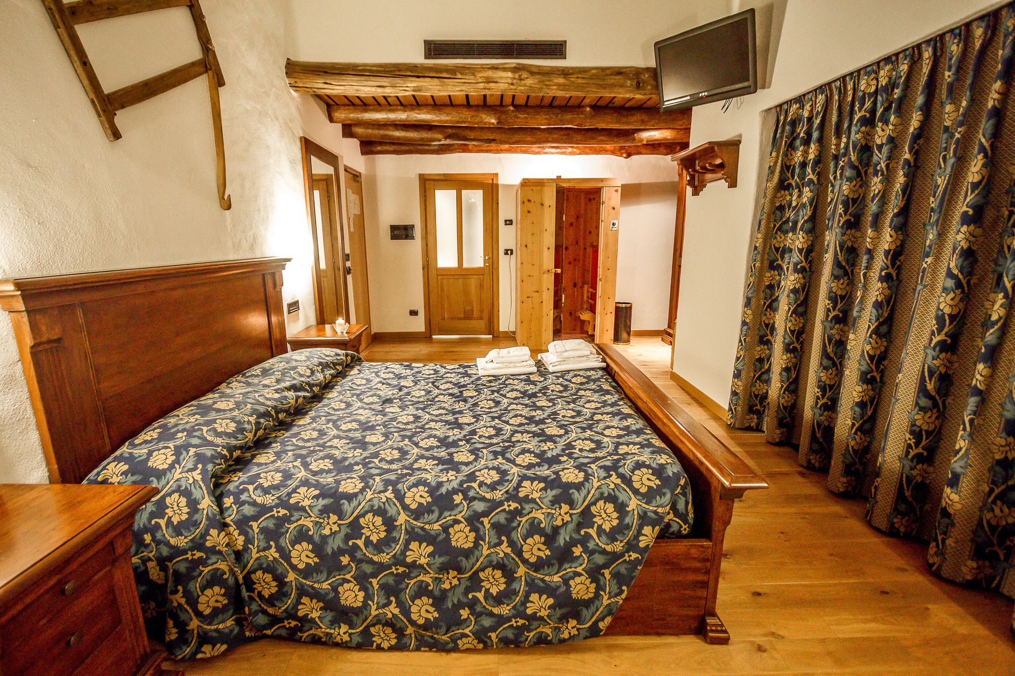 Un dormitorio con cama de madera, una mesita de noche de madera, un televisor montado en la pared y una cortina estampada.