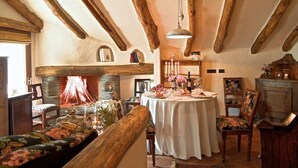 Un interior rústico con vigas de madera a la vista, una chimenea, una mesa redonda lista para una comida y un rincón de estar acogedor.