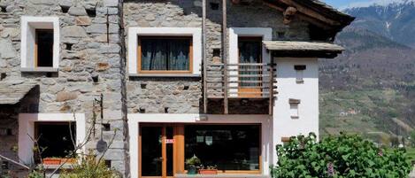 A stone building with a wooden balcony, a glass door entrance, and a view of mountains.