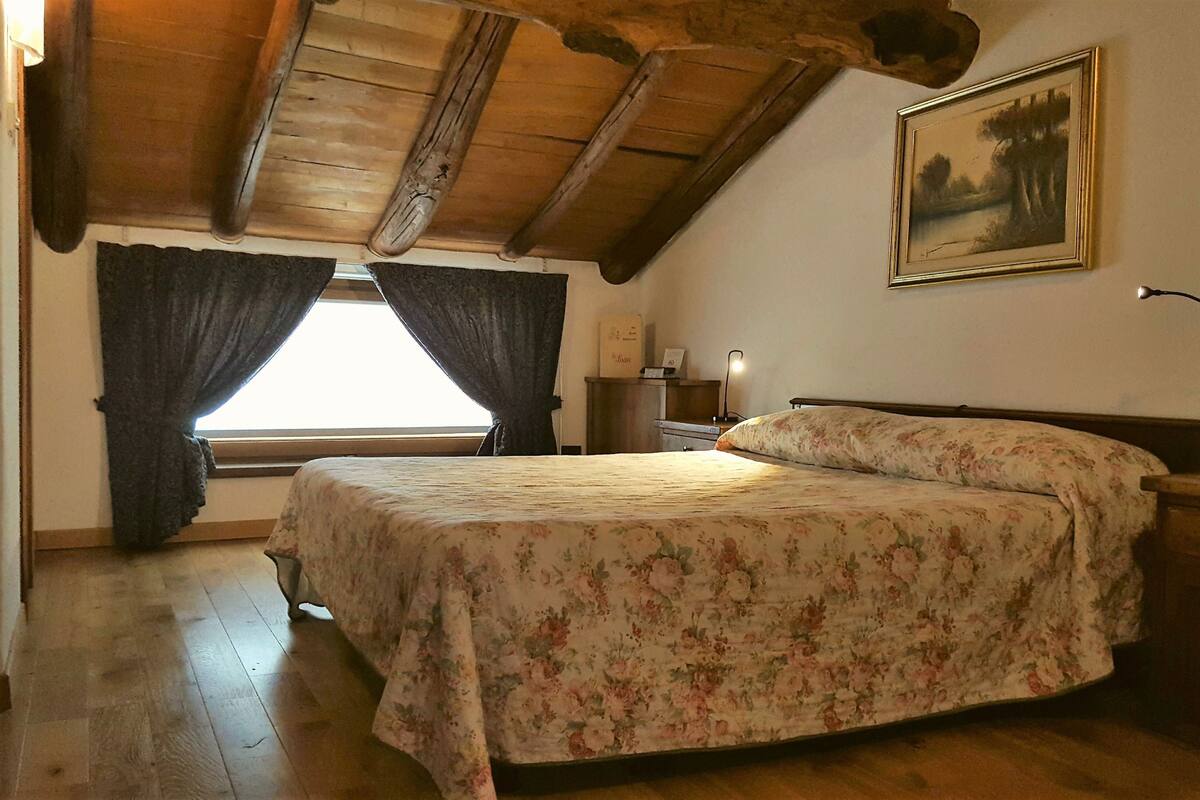Une chambre avec un plafond en bois, un lit recouvert d’une literie à motifs floraux, une table de chevet, une lampe et un tableau accroché au mur.