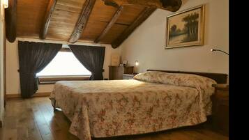 Une chambre avec un plafond en bois, un lit recouvert d’une literie à motifs floraux, une table de chevet, une lampe et un tableau accroché au mur.