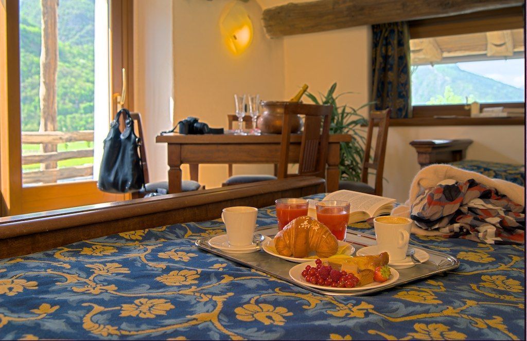 Desayuno con cruasanes, fruta y bebidas sobre una bandeja, una mesa de madera y vistas al exterior con vegetación.
