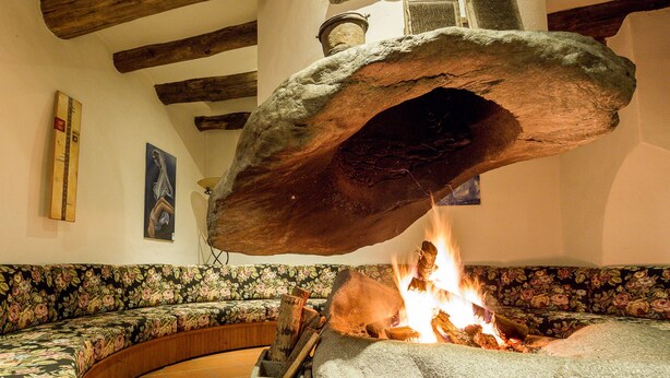 Una chimenea de piedra con fuego encendido, un banco de madera y un sofá estampado con motivos florales.