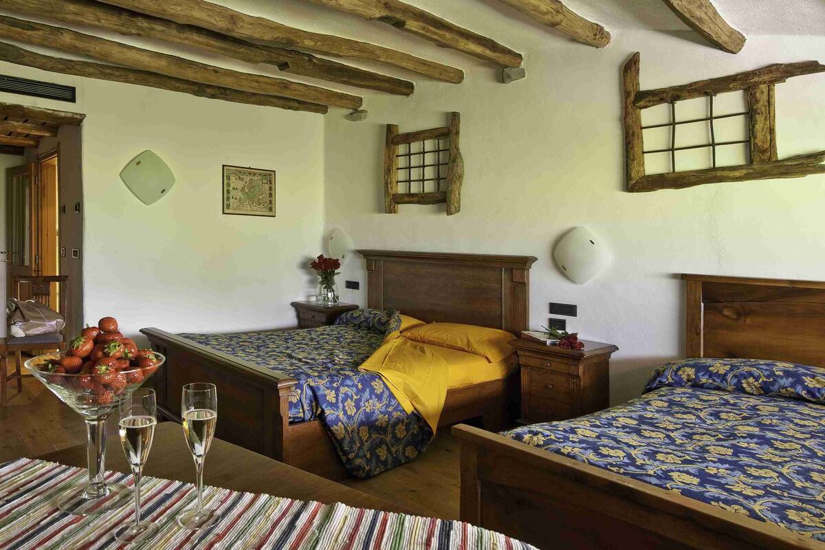 Une chambre avec deux lits, des poutres en bois au plafond, une petite table avec un saladier de fruits et des verres, et un luminaire fixé au mur.