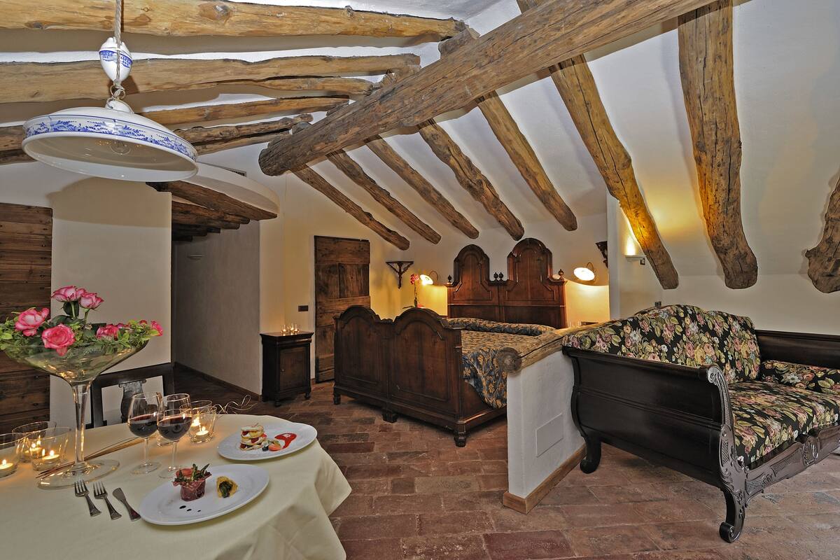 Une pièce rustique avec des poutres apparentes, un lit en bois, un canapé à motifs floraux et une table à manger dressée avec des assiettes et des verres.