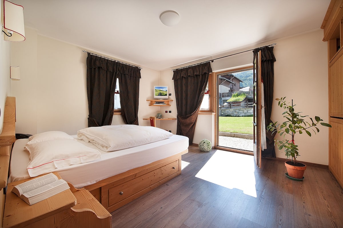 A bedroom with a wooden bed, a small bedside table, a potted plant, and a view of the outdoors.