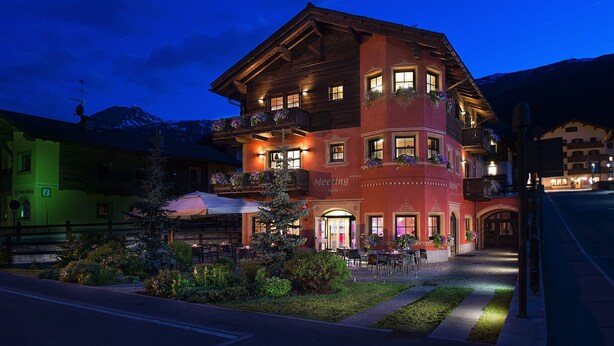 A hotel with a red facade, multiple balconies with flower boxes, and a well-lit outdoor seating area. - Hotel Meeting (Livigno)
