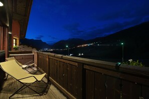 A balcony with wooden railings and a lounge chair, overlooking a mountainous landscape with lights in the distance.