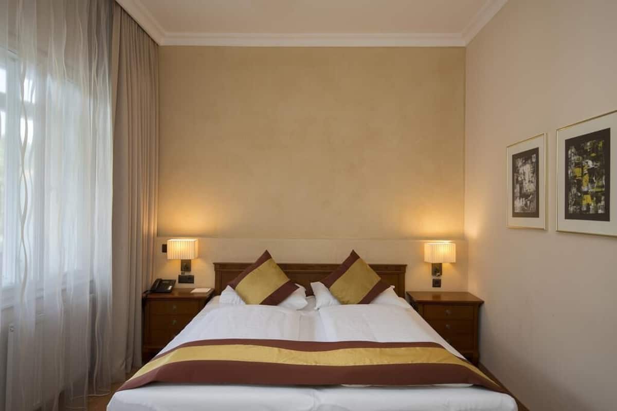 Ein Hotelzimmer mit einem großen Bett, zwei Nachttischen mit Lampen, einem Fenster mit durchsichtigen Vorhängen und zwei gerahmten Bildern an der Wand.
