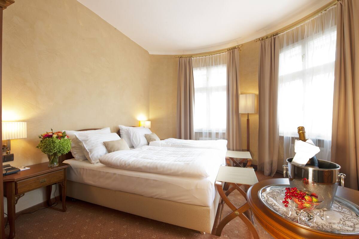 Ein Hotelzimmer mit einem großen Bett, zwei Nachttischen, einem Schreibtisch und einem Stuhl. Auf dem Nachttisch steht eine Vase mit Blumen und auf dem Schreibtisch ein Tablett mit Früchten und einer Flasche.