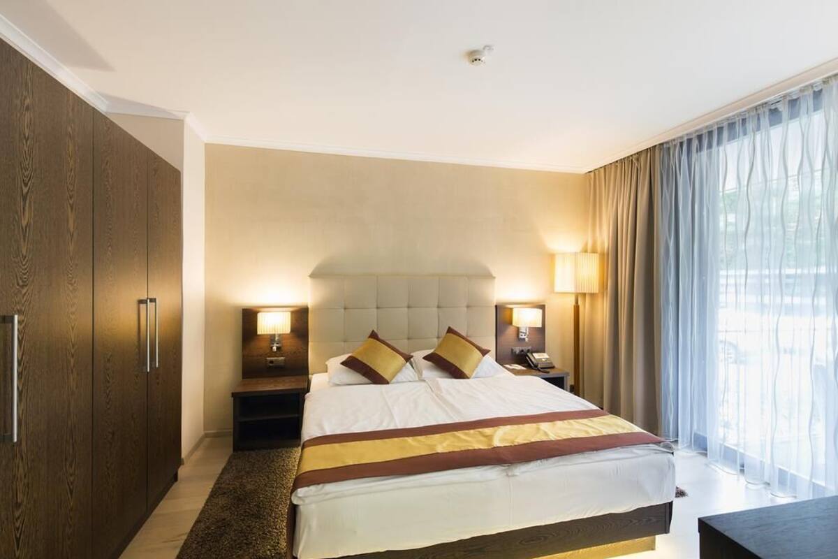 Ein Hotelzimmer mit einem großen Bett, zwei Nachttischlampen, einem Holzschrank und Blick ins Freie durch durchscheinende Vorhänge.