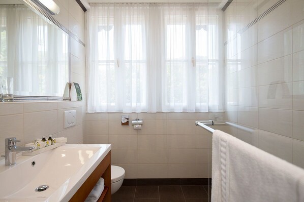 A modern bathroom with a white sink, toilet, and bathtub. There are two towel racks and a window with sheer curtains.