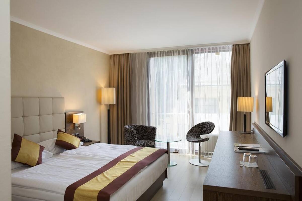 Ein Hotelzimmer mit einem großen Bett, zwei Sesseln, einem Schreibtisch und einem Fernseher an der Wand.