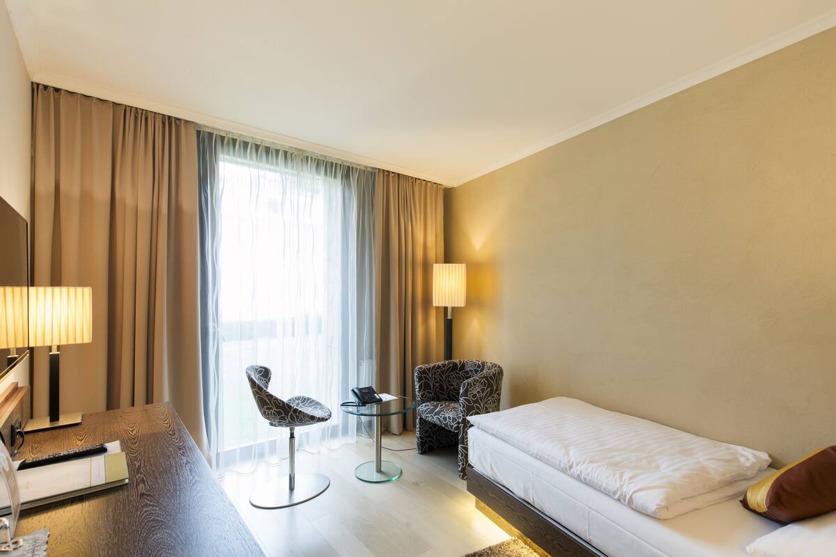 Ein Hotelzimmer mit einem Bett, einem Schreibtisch mit Lampe, einem Sessel und einem Fenster mit Vorhängen.