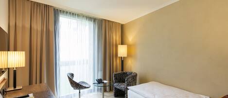 Una habitación de hotel con una cama, un escritorio con lámpara, una silla y un ventanal con cortinas.