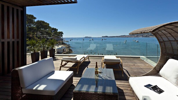 A deck with white cushioned seating, a glass-top table, and a bottle of wine. - Hostellerie La Farandole (Sanary-sur-Mer)