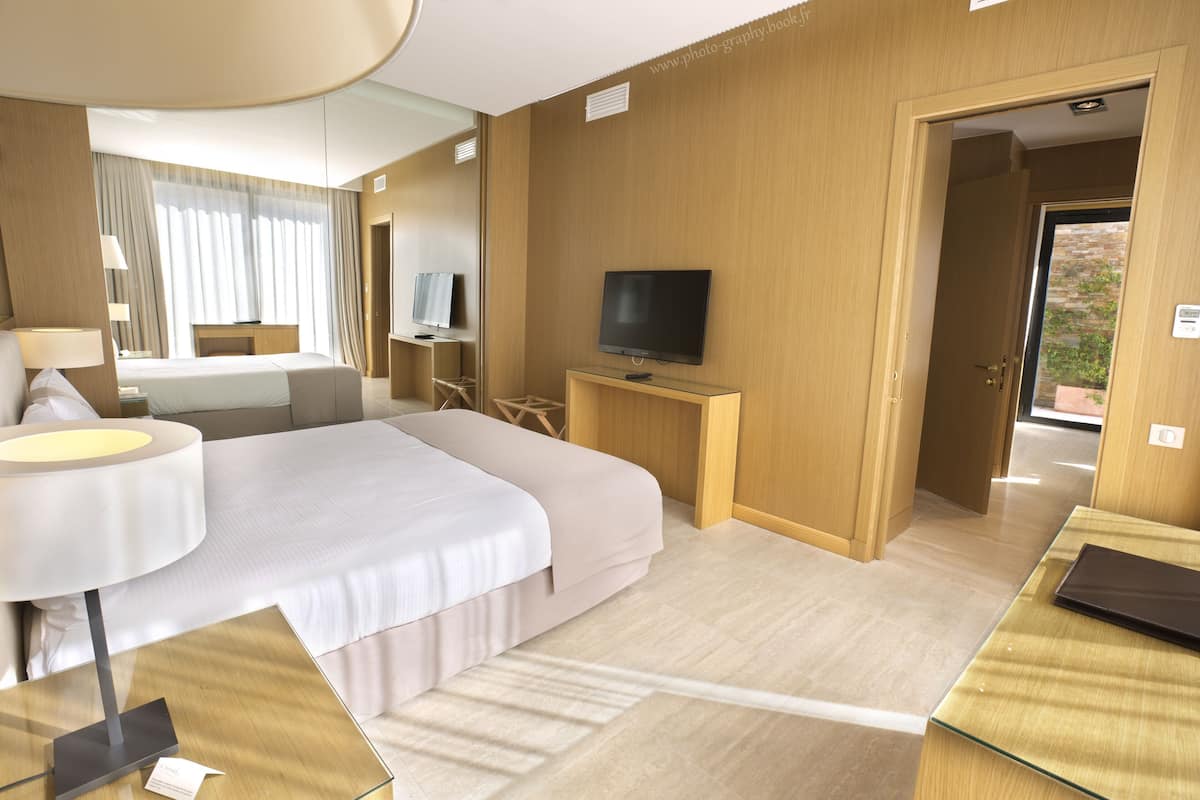 Habitación de hotel con una cama grande, un televisor sobre un mueble de madera, un escritorio con una computadora portátil y una puerta que da a un espacio exterior.