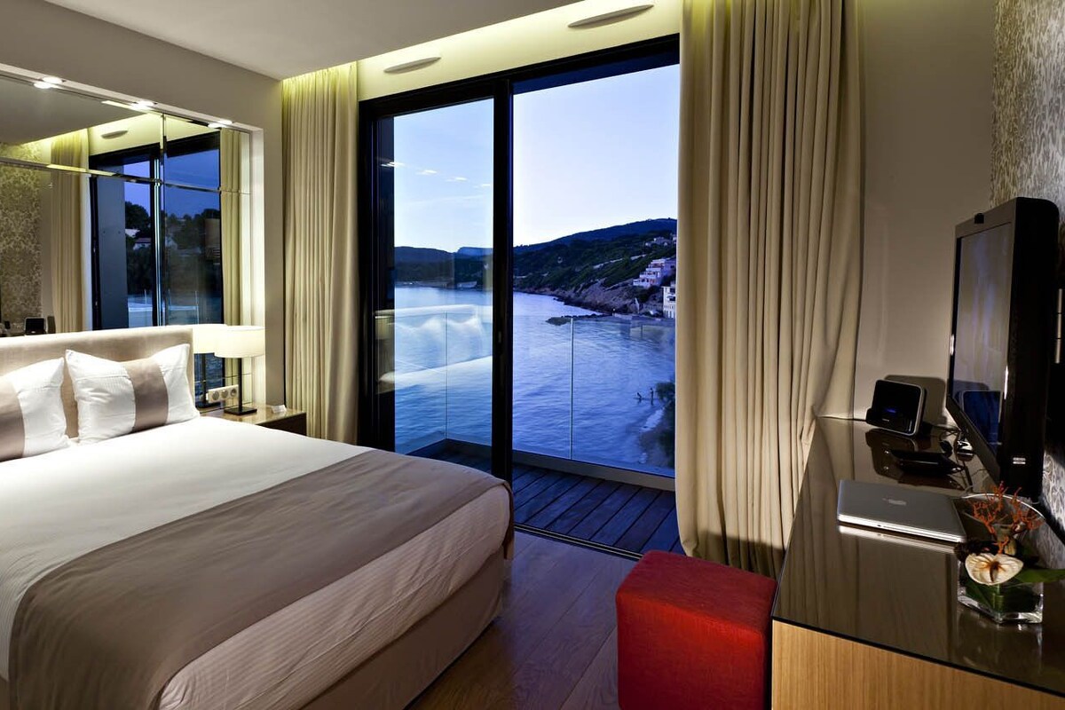 Una habitación de hotel moderna con una cama grande, televisión, escritorio con una computadora portátil y vista al mar.