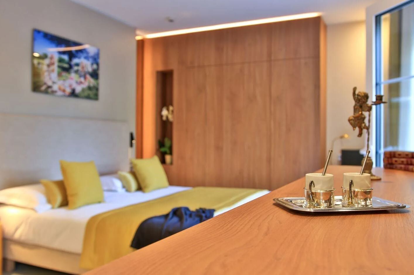 A hotel room with a bed, a wooden headboard, a nightstand with a tray of glasses, and a large window.